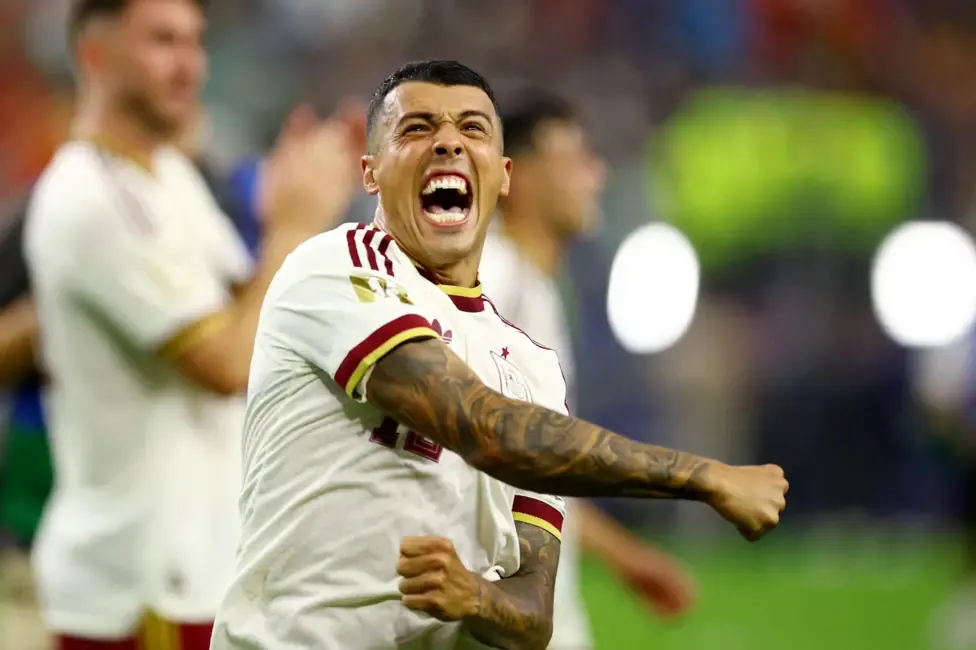 A male footballer shouts in joy while flinging his right arm across his chest in celebration. He is wearing a white shirt with red and yellow trim and standing in a football stadium with green grass. Other players can be seen out of focus behind him.