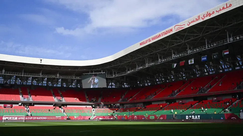 A general view of the inside of the Moulay Hassan Stadium in Rabat, looking towards one corner, showing red and green seats. The green pitch is partly in shadow as a blue sky with light cloud shines above the grey roof which arcs around and contains the name of the stadium in red English and Arabic text
