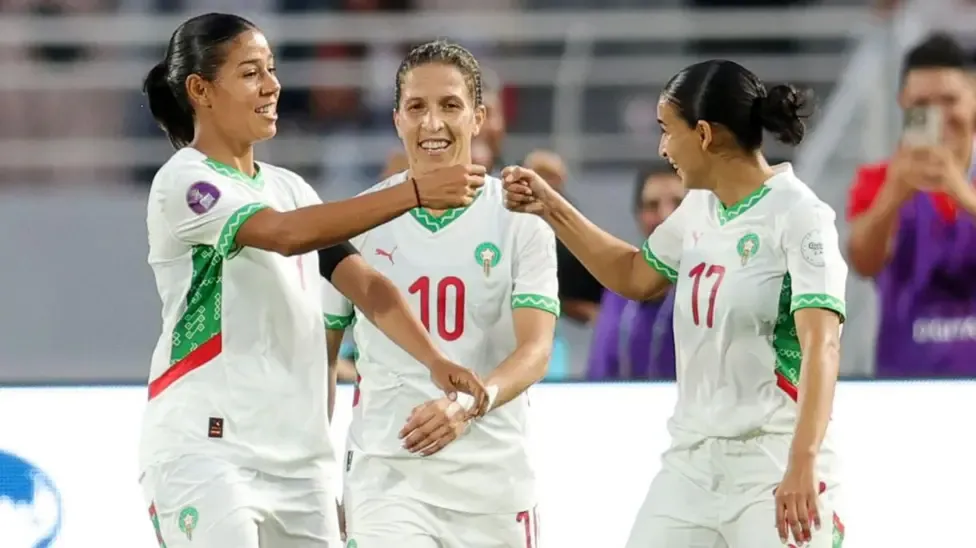 Ghizlane Chebbak (left) celebrates with two of her Morocco team-mates by going to do a fist bump with one of them. All three are smiling and wearing all-white kit with red and green trim. A man in a purple tabard can be seen blurred in the background holding up a phone with two hands to take a picture